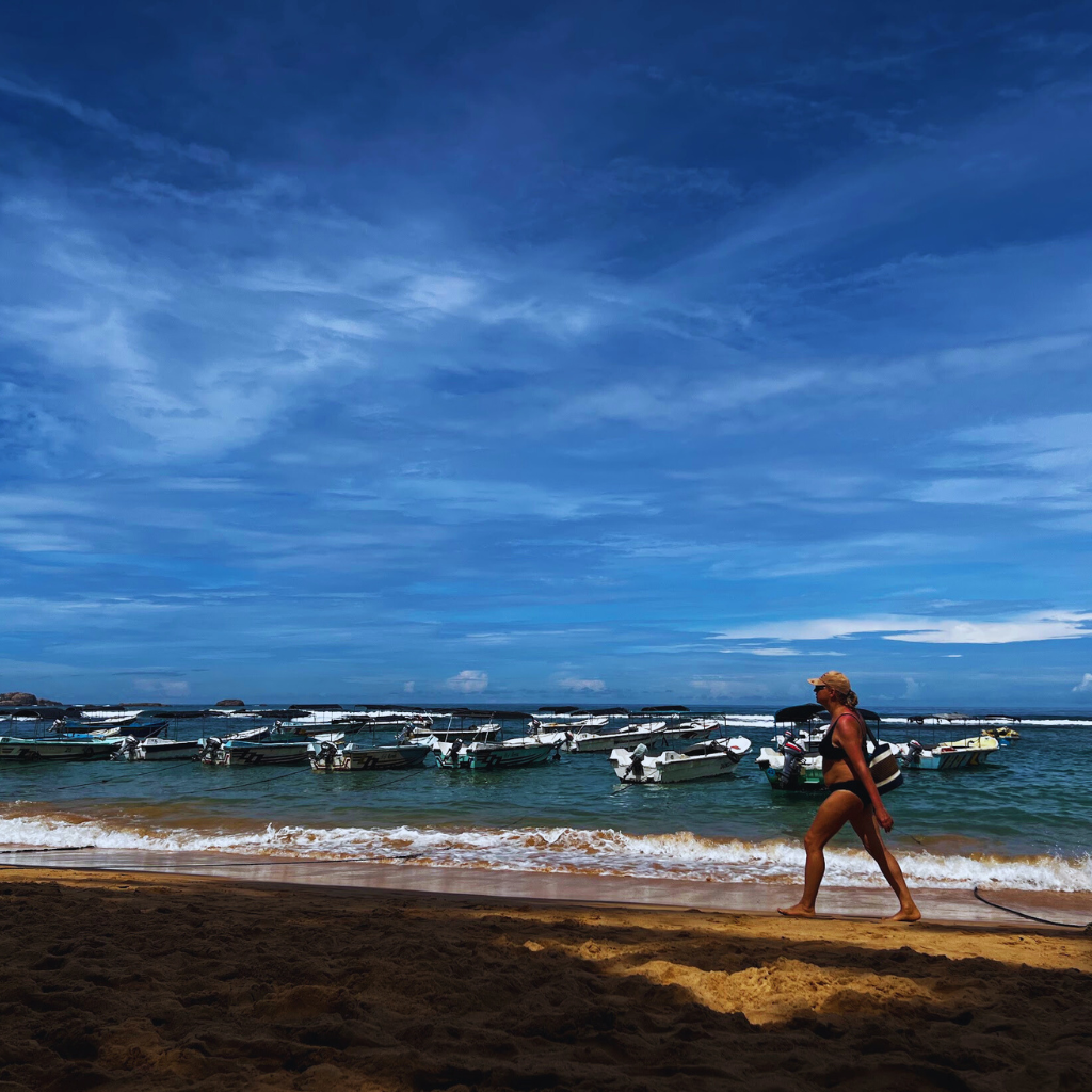 Person walking along a beach with boats anchored in the water under a clear blue sky in Sri Lanka